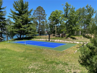 Outdoor residential sports court with a blue and green surface, a basketball hoop, and a tennis net, surrounded by trees and near a lake in Eau Claire.