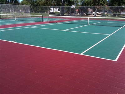 Outdoor sports court with a green and red surface, featuring multiple tennis nets and surrounded by a fence and trees in Brookfield.