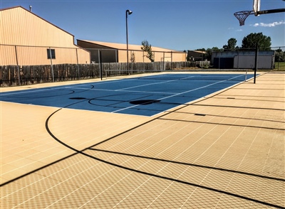 Outdoor sports court with a blue and beige surface, featuring basketball hoops and tennis nets, surrounded by a fence.